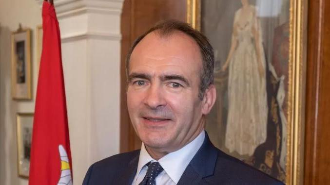 Alfred Cannan smiling. He is wearing a dark suit and tie with a white shirt, and is standing next to a Manx flag. There is a portrait of Queen Elizabeth II on the wall behind him.