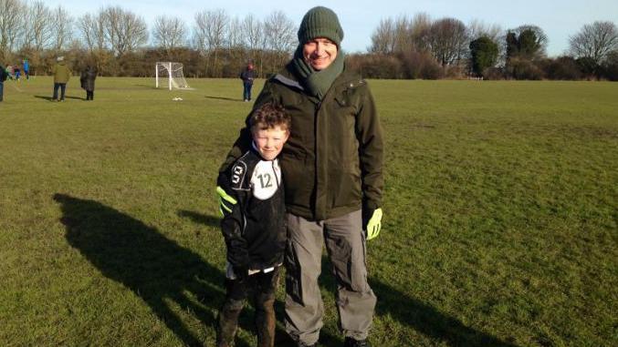 A picture of a young boy in a rather muddy dark strip on a football pitch with his father who has his arm around him.