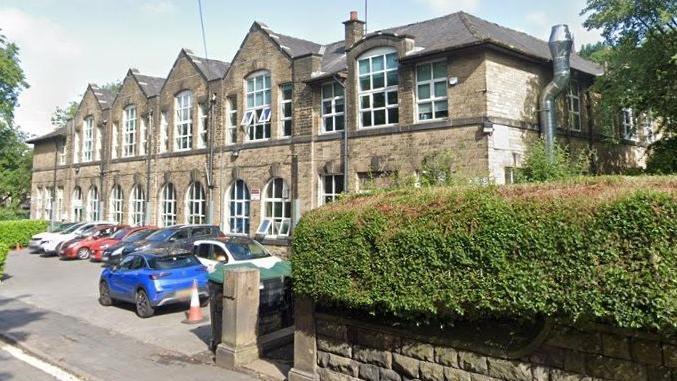 Castle Hill Primary School, Todmorden, a building from 1912 with two storeys, a hedge in front and several parked cars. 