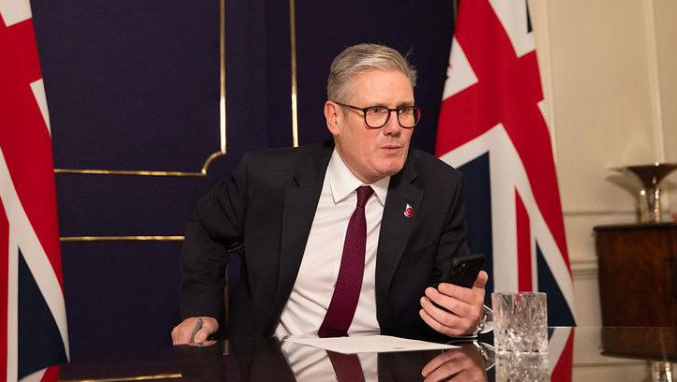 Sir Keir Starmer holsd his mobile phone in front of him as he speaks to Donald Trump from his office in No 10 Downing Street. There are two union jack flags behind him.