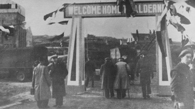 A black and white image of people returning to Alderney. The walk under a sign saying 'Welcome home to Alderney'