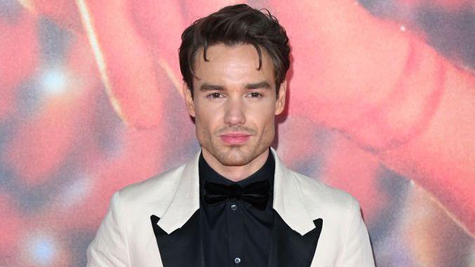 Liam Payne has brown hair and is wearing a white blazer and a black shirt. He is standing in front of a red background with a slight smile on his face. 