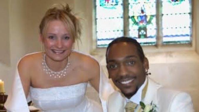 Karin Burke in a white wedding dress next to Richard who is wearing a white suit with a gold tie and waistcoat. The pair are in front of a stone wall of a church with a stained glass window behind them.