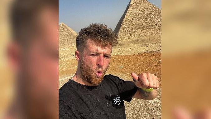 Tommy on a run in the desert with two pyramids in the background. He has short ginger hair and a beard and is doing a thumbs up sign at the camera.