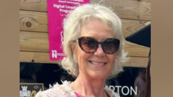 Liz is standing in the middle of the picture wearing big sunglasses and smiling. She is wearing a pink top and a light pink cardigan. 