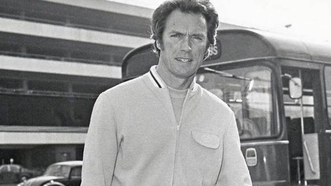 Clint Eastwood in a bomber jacket looks at the camera as a bus passes behind