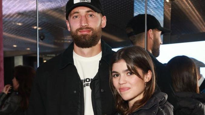 A man wearing a cap stands beside a woman wearing a black coat