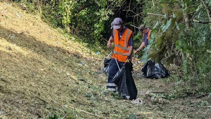 Men in orange high-visibility jackets clearing rubbish in a grassy area using litter pickers and bin bags