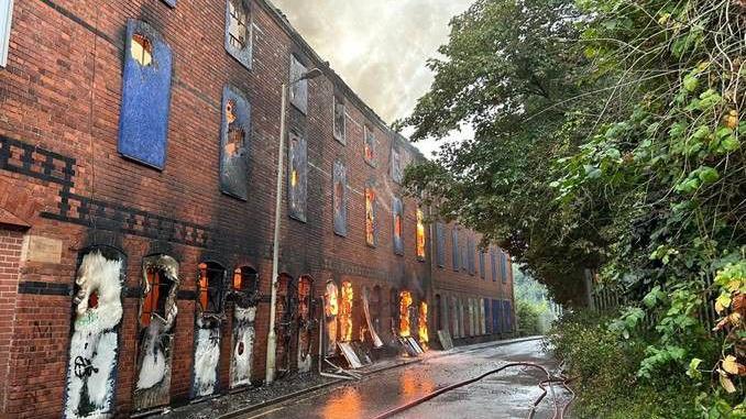 A three-storey derelict red-brick building with boarded up windows. Flames can be seen through the windows of the first two storeys and fire hoses are running down the street.