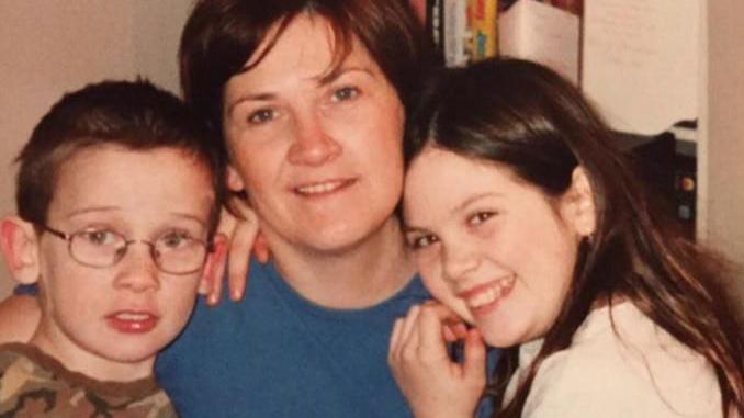 Josh Hanson as a child with short dark hair and silver-framed glasses. He is wearing a brown T-shirt. He is standing next to his mother who has long dark hair and is wearing a blue top. To her left is Brooke, with long dark brown hair, wearing a white top.