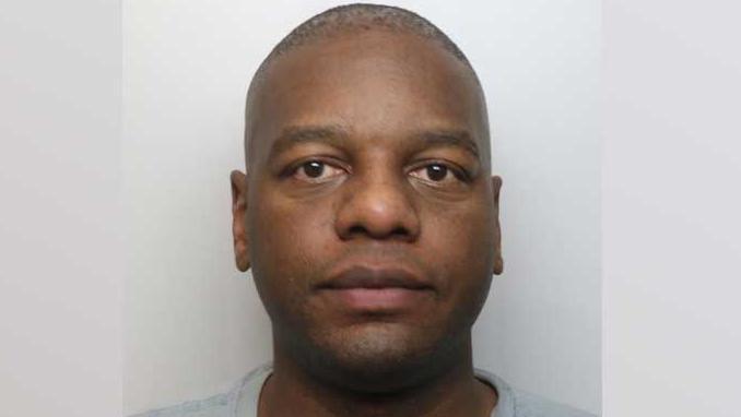 Mugshot of John Malonda, who has close-cropped hair, large brown eyes and is wearing a grey prison jumper.