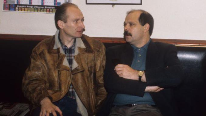 Billy Hutchinson and David Ervine sitting in a bar.  David Ervine is on the right, and he is wearing a black jacket and blue shirt, he has a moustache and brown hair. Billy Hutchinson is wearing a blue and red check shirt with a brown and white jacket. He has brown hair