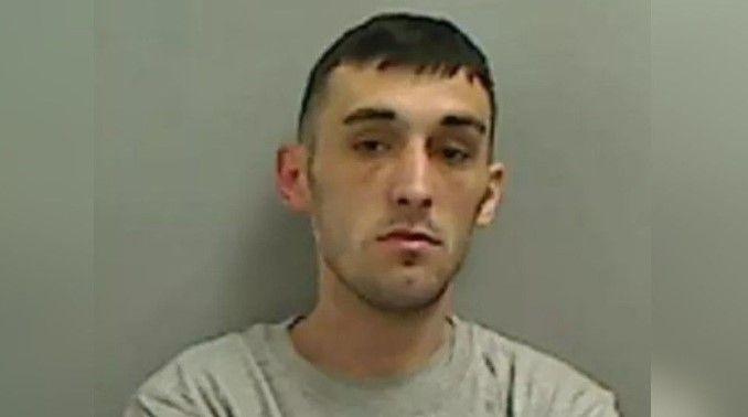 Liam Matthews' police mugshot, he is white, thin and clean-shaven with short dark hair. he is looking at the camera with no expression and is wearing a grey-coloured crew-neck top
