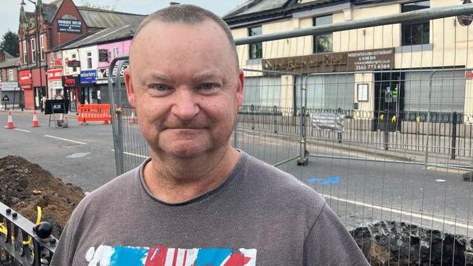 Jonathan Higgin has a shaved head, a grey T-shirt with a Union flag on it, and is standing in front of the hole.