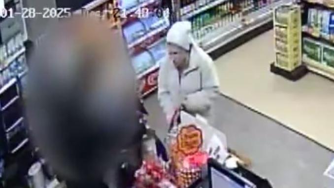 CCTV footage of Julie Buckley in a Budgens shop in March. She is wearing a white wooley hat and white coat and has a shopping bag in her left hand. The worker serving her is blurred out. 