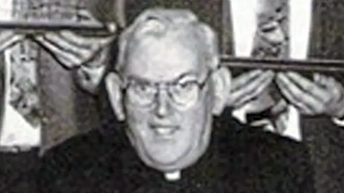 A black and white photo of the late Fr Malachy Finegan.  He had short white hair and glasses and was wearing a black suit and a priest's collar.