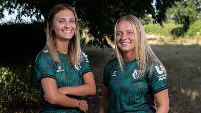 Two young women stand in the countryside on a grass track under some large trees. They wear the new Watford away shirt which is a dark forest green with lighter green checks. They smile at the camera and both have long straight fair hair. It is a sunny day.