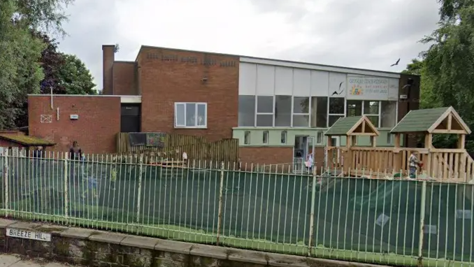 An exterior shot of the Early Learners Day Nursery, a brick building with a flat roof. There is a playground in front of the building including a wooden structure with slide attached. The playground is separated from the street by a low stone wall topped with railings.
