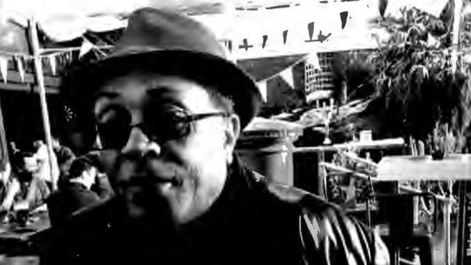 Louis Campbell is wearing a black leather jacket, black polo top and a black hat. he has black sunglasses and is sat in a busy pub garden