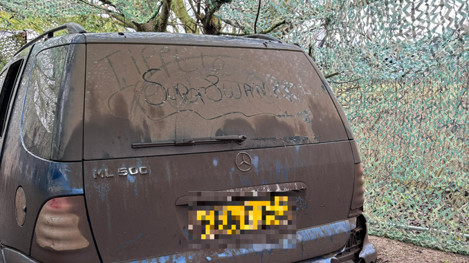 A dirty blue car under some green netting. The registration plate has been pixelated and a word - which cannot be made out - has been written on the rear view window. 