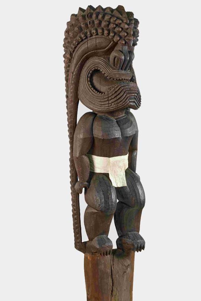A wooden sculpture of a human-like figure wearing a large helmet that covers the face and extends at the back to the floor. Is it wearing a white loin cloth. 