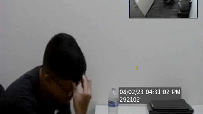 A man is seen holding his head in his hands at a desk in an interrogation room. A bottle of water is on the desk and the photo is blurry. In the top right corner a smaller inserted photo shows the entirety of the room from above with two FBI agents.