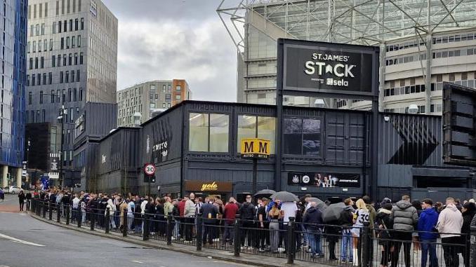 A queue of people standing outside St James' Stack which is made up of black shipping containers.