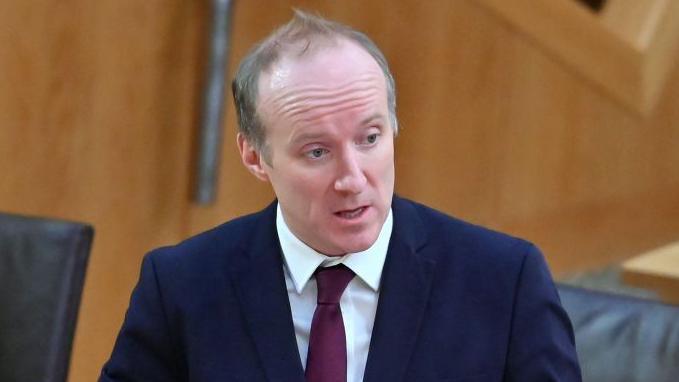 Michael Marra in a dark blue suit and burgundy tie stands and speaks in a chamber with wooden desks and black chairs visible around them.
