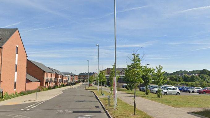 A long residential road with houses on the left, and car parking on the right. There are lots of trees and grass areas surrounding the car park