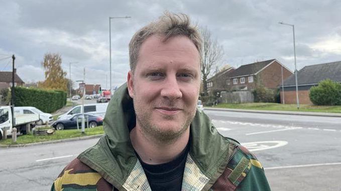 A blonde man with short hair and stubble. He is wearing a camouflage jacket.