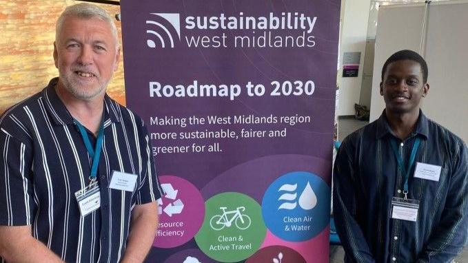 Two men pose either side of a banner stating sustainability West Midlands, Roadmap to 2030. The men are both wearing striped shirts and lanyards. The one on the left has grey hair and a beard and the other has close cropped black hair.