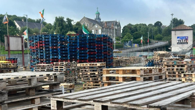 Shows large piles of pallets being stacked in preparation for a bonfire. Flags, including the Irish tricolour and Palestinian flag, are seen in the background.