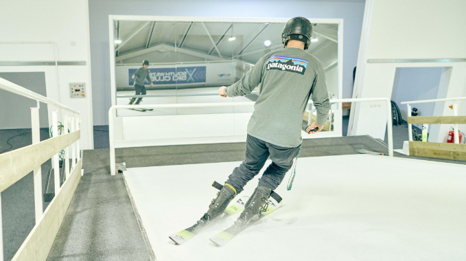 Someone using the ski slope at the site. They are wearing a brown top and ski trousers and can see themselves in a large mirror screen