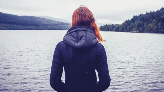 Young woman with red hair and a black hooded top, her back to the camera and her hands in her pockets, looks out at a lake