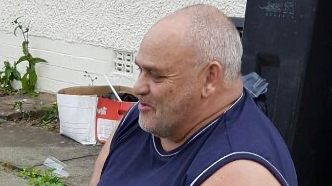 A man with short white hair and a dark blue vest looking off camera while sitting in front of a white wall.