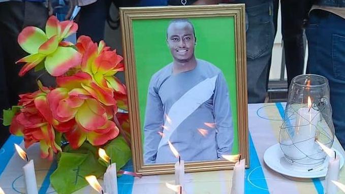 A frame with a photo of a smiling Francis Njuki, with candles in front, at his burial in his village in Kenya