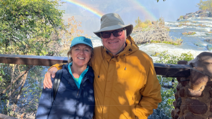 The couple are stood by a waterfall with rainbows appearing behind them as they stand against a fence. Mr Cherry has a hat on and a yellow jacket and is smiling with his arm around his wife who has a blue cap on and blue jacket and is also smiling.