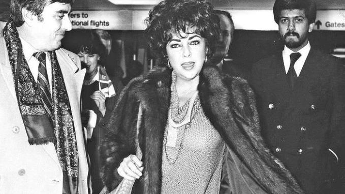 Elizabeth Taylor wearing a fur coat and dark eye makeup is surrounded by people are she arrives at Heathrow Airport