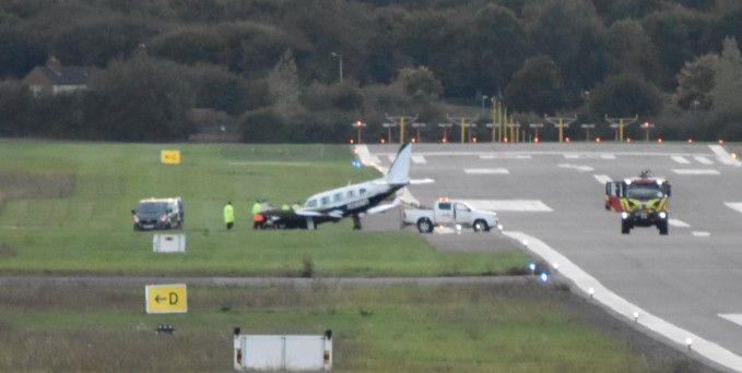 A picture of the runway at Gloucestershire Airport. The plane can been seen on the grass, with the nose slightly pointed down. At least two vehicles appear to be next to the plane. 