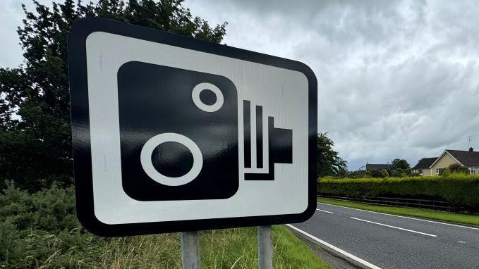 A black and white speed camera ahead sign mounted on a grass verge at the side of a single lane asphalt carriageway.