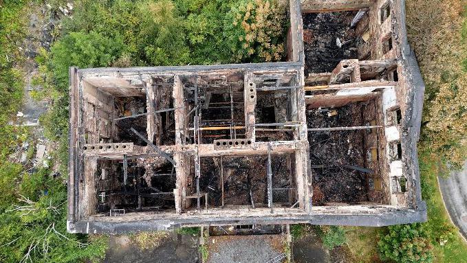 A drone image shows the burned-out shell of Woolton Hall directly from above. The roof is completely gone and only the walls of the building remain.