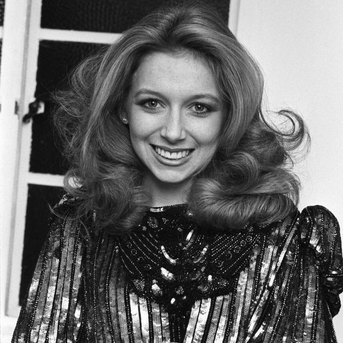 An older Lena Zavaroni, aged 21, smiles at the camera, with a big blowout hairstyle. She is wearing a metallic sequin dress. The photo is in black and white.