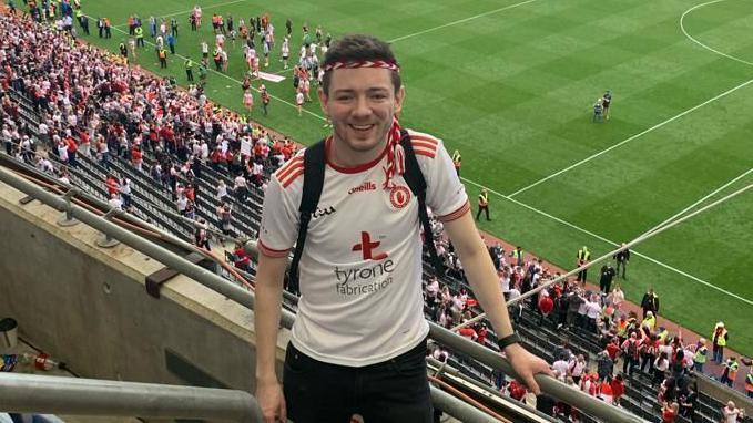 Cahir wearing Tyrone white GAA top with red and white rope around his head. He is standing high in the stands at Croke Park with ones gathered on the pitch and stand behind him