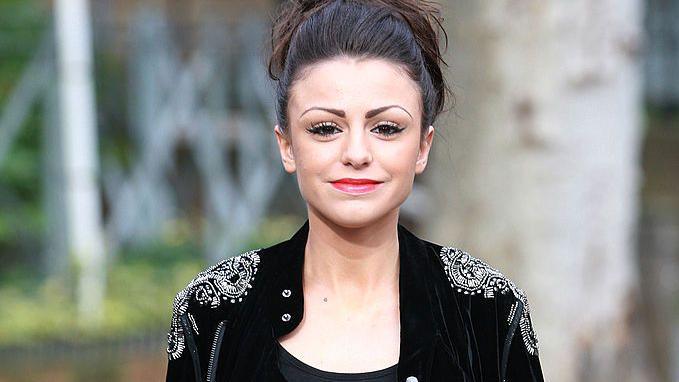 A young woman pictured from the shoulders up as she walks down the street. She's got a neutral expression, and wears a black blazer with intricate white patterns embroidered into both shoulders. Her black hair is loosely tied up.