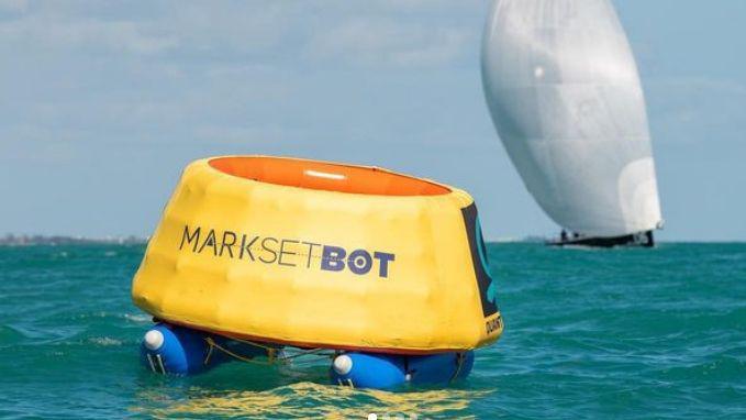 A MarkSetBot, a round yellow circular robot about 1.5m-tall on a catamaran-like float,  on the sea, with a yacht behind it