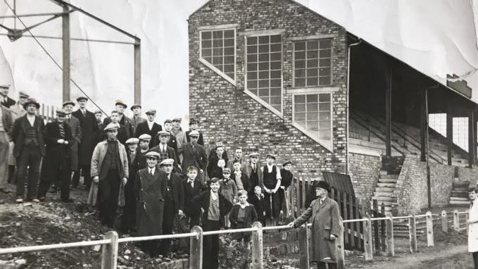 A black and white image of fans standing next to the stand at Borough Park in 1937, The stand is a brick construction with steps at the front leading up to the seats. The fans are standing on a mud bank. Their are a few boys near the front while the rest are men all wearing hats. 