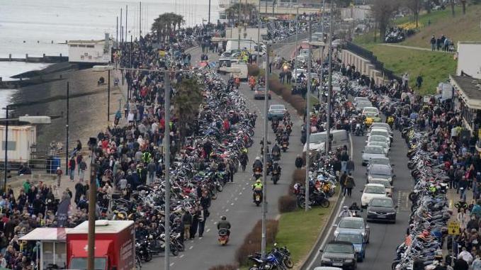 Hundreds, if not thousands, of motorcycles and people lining a seafront road as more motorcycles drive along it.