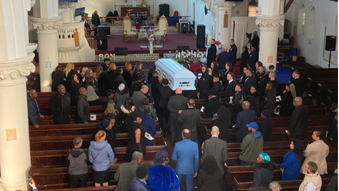 Mr Meade's white coffin is brought in to the church with mourners behind and standing in the pews.