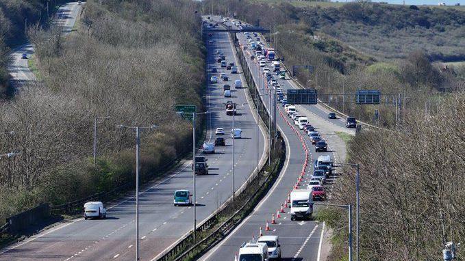 A two carriage A road. The carriageway on the left has one lane closed by cones and congested traffic.
