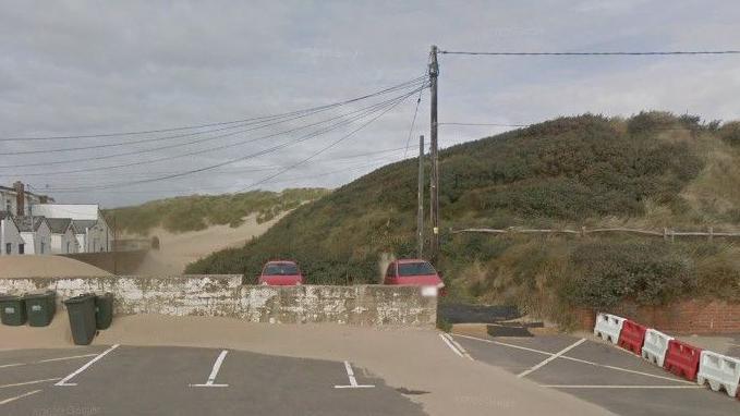 A Google maps image showing high sand dunes covered in greenery on the outskirts of a beachside carpark. Small white cottages can be seen in the distance on the left.